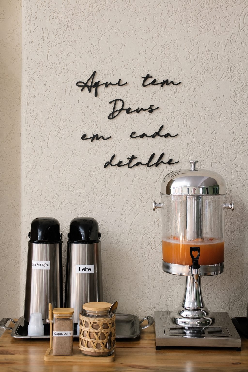 Parede do salão do café com os dizeres Aqui tem Deus em cada detalhe, garrafas térmicas e suqueira
